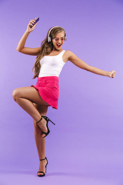 Full length portrait of happy energetic woman 20s in summer clothes and sunglasses dancing while listening to music via wireless headphones isolated over violet background - Fotoğraf, Görsel