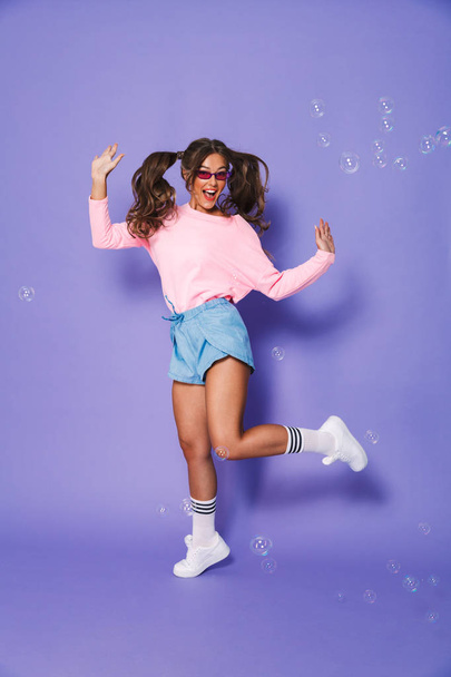 Full length portrait of european teenage girl with two ponytails in sweatshirt and sunglasses laughing while having fun isolated over violet background in studio - Foto, Bild