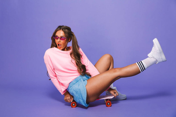 Portrait of a cute girl in sweatshirt posing on skateboard isolated over violet background - Photo, Image