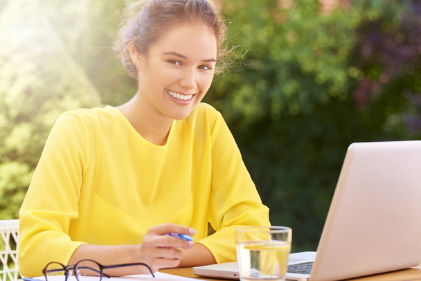 Portrait of young smiling woman using her laptop and writing something sitting outdoors. - Photo, Image