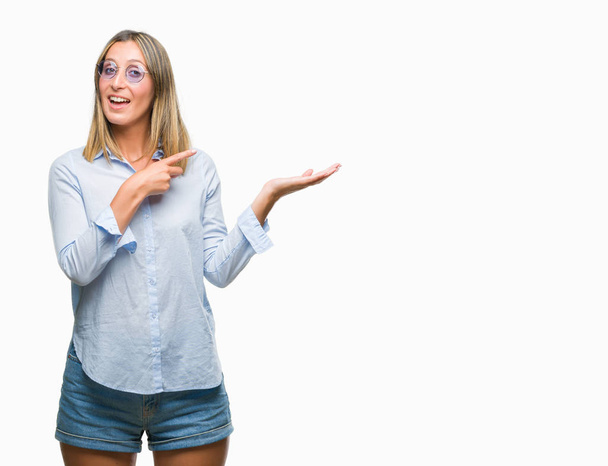 Mooie jongedame dragen van een zonnebril op geïsoleerde achtergrond verbaasd en lachend naar de camera tijdens het presenteren met hand en met de vinger te wijzen. - Foto, afbeelding