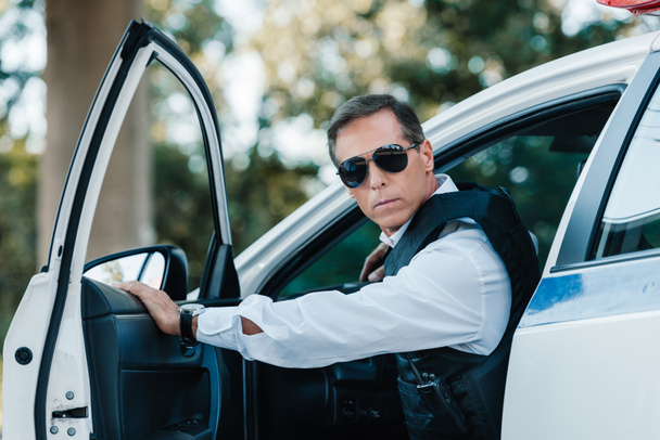 selective focus of mature policeman in bulletproof vest and sunglasses walking out from car  - Foto, immagini