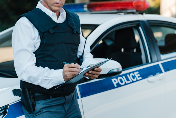 частковий вид на чоловіка середнього віку поліцейського в куленепробивному жилеті, що пише в буфері біля автомобіля на вулиці
  - Фото, зображення