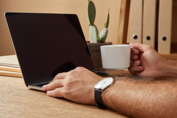 imagem cortada do homem usando laptop com tela em branco e segurando xícara de café na mesa
  - Foto, Imagem