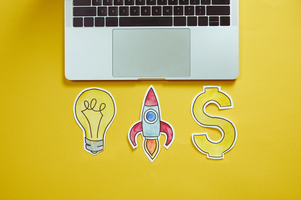 top view of laptop, paper lightbulb, paper rocket and dollar sign on yellow surface - Foto, imagen