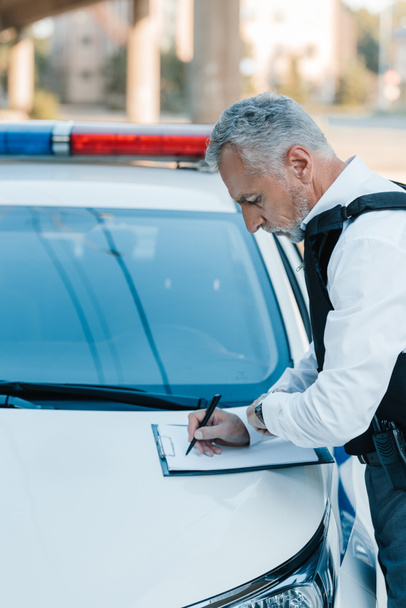 side view of focused male police officer writing in clipboard on car at street - Photo, Image