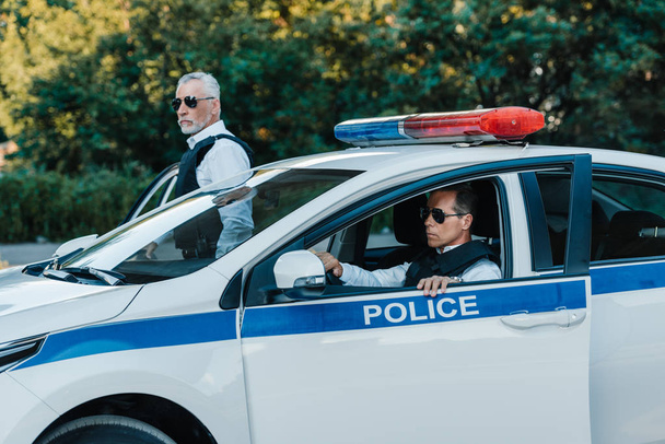 middle aged policemen in sunglasses walking out from car at urban street  - Photo, Image