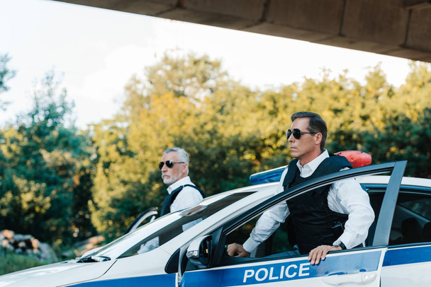Selektivní fokus starší policista v sluneční brýle a neprůstřelné vesty, stojící vedle auta na ulici  - Fotografie, Obrázek