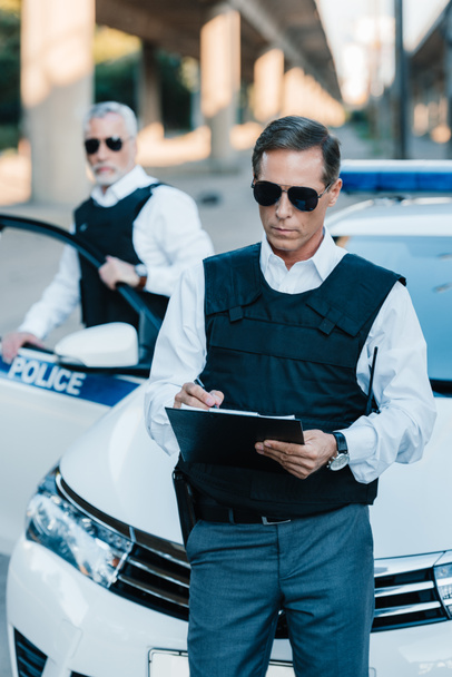 male police officer in sunglasses writing in clipboard while his colleague standing near car at street - Fotografie, Obrázek