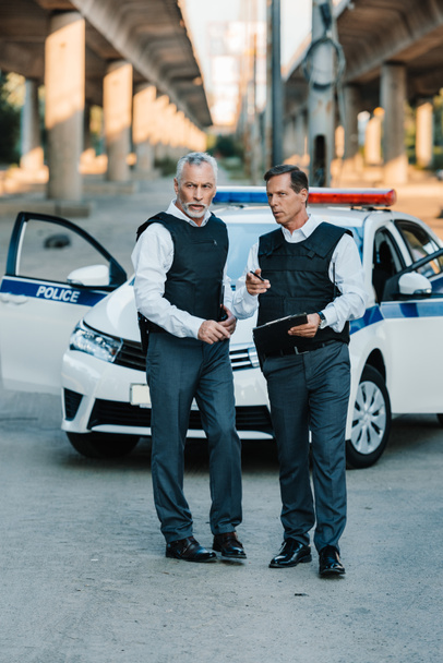мужчина-полицейский с планшетом, указывающим пальцем на коллегу, стоящего рядом с машиной на улице
 - Фото, изображение