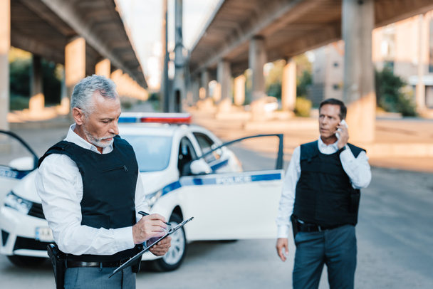 Polizist mittleren Alters schreibt in Klemmbrett, während sein Kollege auf der Straße mit dem Smartphone spricht  - Foto, Bild