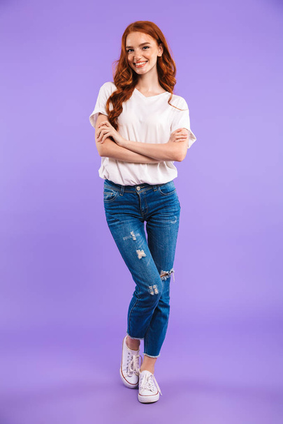 Full length portrait of a smiling young girl standing isolated over violet background, arms folded - Foto, imagen
