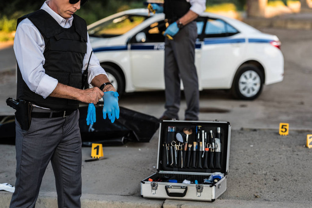 częściowy widok mężczyzna policjant w okulary zakładanie rękawic lateksowych, podczas gdy jego kolega stojący na zbrodni z trupem w ciało torba  - Zdjęcie, obraz