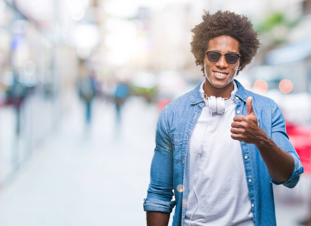 Afro american człowiek noszenia słuchawek, słuchanie muzyki na na białym tle robi szczęśliwy kciuk w górę gest ręką. Zatwierdzająca wyrażenie patrząc na kamery z pokazując sukces. - Zdjęcie, obraz