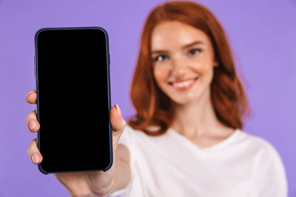 Portrait of a happy young girl standing isolated over violet background, showing blank mobile phone - Fotó, kép