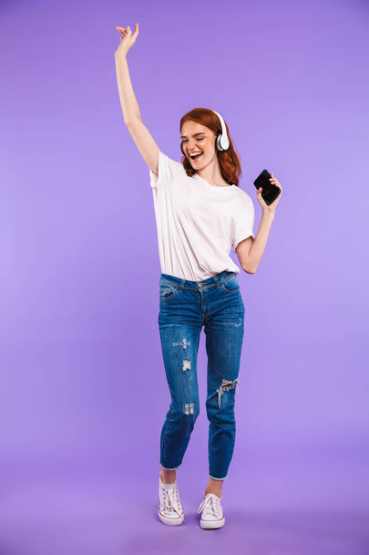 Full length portrait of a smiling young girl standing isolated over violet background, holding mobile phone, listening to music with headphones, dancing - Zdjęcie, obraz