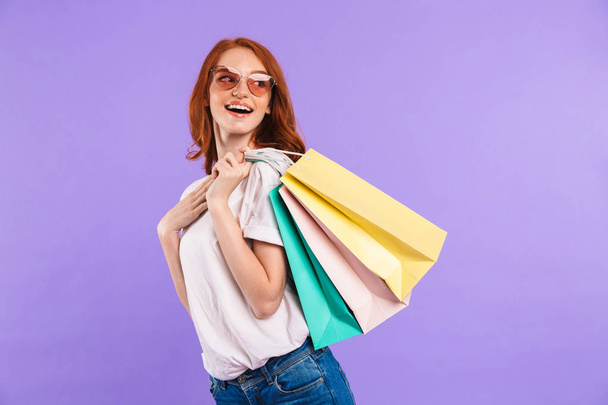 Portrait of a cheerful young girl in sunglasses standing isolated over violet background, carrying shopping bags, looking away - Photo, Image
