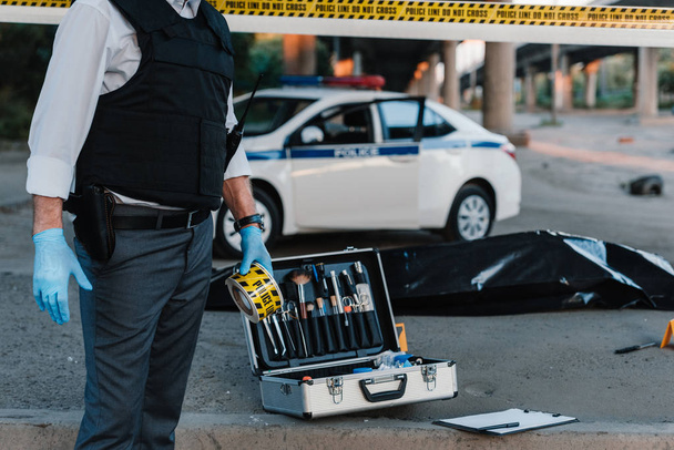 vue partielle du policier en gants de latex tenant la ligne de police sur les lieux du crime avec cadavre dans un sac mortuaire
  - Photo, image