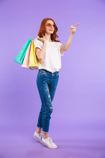 Full length portrait of a smiling young girl in sunglasses standing isolated over violet background, carrying shopping bags, pointing away - Foto, afbeelding