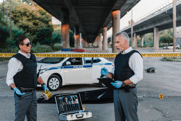 policier mature avec presse-papiers parlant à un collègue dans des lunettes de soleil près du corps dans un sac mortuaire sur les lieux du crime
 - Photo, image