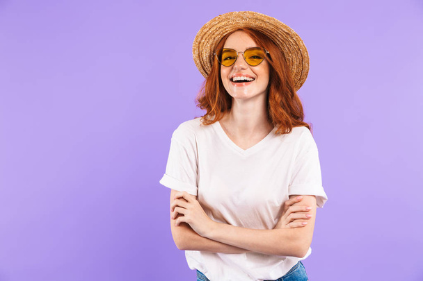 Portrait of a cheerful young girl in straw hat and sunglasses standing isolated over violet background - Foto, afbeelding