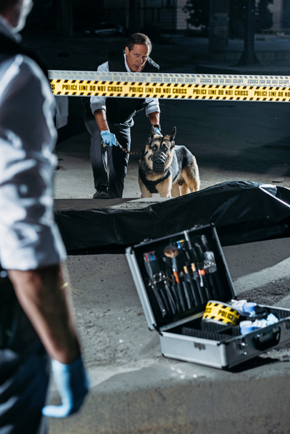 partial view of police officer standing near case with investigation tools while his colleague standing with german shepherd on leash near corpse at crime scene  - Photo, Image