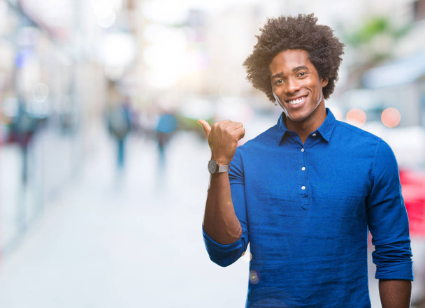 Hombre afroamericano sobre fondo aislado sonriendo con cara feliz mirando y señalando hacia un lado con el pulgar hacia arriba
. - Foto, imagen