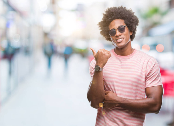 Афро-американец в солнечных очках на изолированном фоне улыбается со счастливым лицом, глядя и указывая на сторону с большим пальцем вверх
. - Фото, изображение
