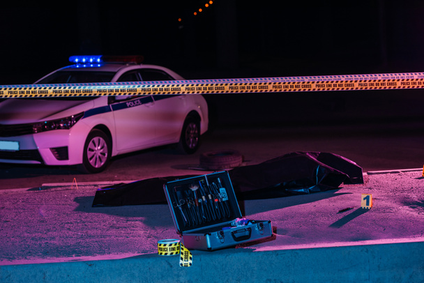 toned picture of crime scene with police car, case with investigation tools, cross line and corpse in body bag - Photo, Image