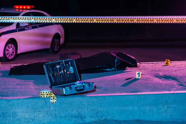 foto tonificada do caso com ferramentas de investigação, linha cruzada, carro da polícia e cadáver em saco de corpo na cena do crime
  - Foto, Imagem
