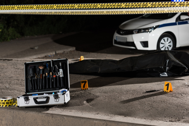 case with investigation tools, car, police line and corpse in body bag at crime scene  - Photo, image