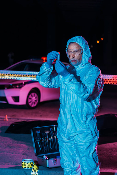 focused male criminologist in protective suit and latex gloves collecting evidence near cross line at crime scene  - Foto, Imagen