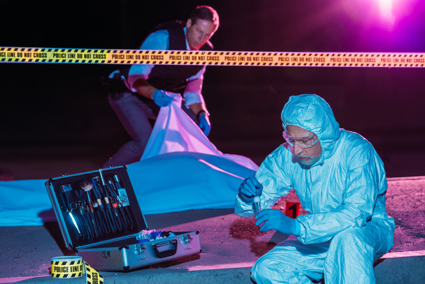 criminólogo masculino de mediana edad recogiendo pruebas mientras el policía cubre el cadáver en la escena del crimen
  - Foto, Imagen