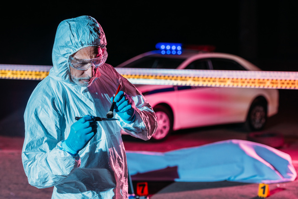 concentrado maduro masculino criminologista em terno protetor e luvas de látex tomando impressões digitais de faca na cena do crime
  - Foto, Imagem
