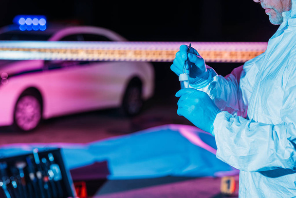 防護服と死体を犯罪現場でピンセットでフラスコに証拠を入れてラテックス手袋で男性の犯罪の部分的なビュー  - 写真・画像