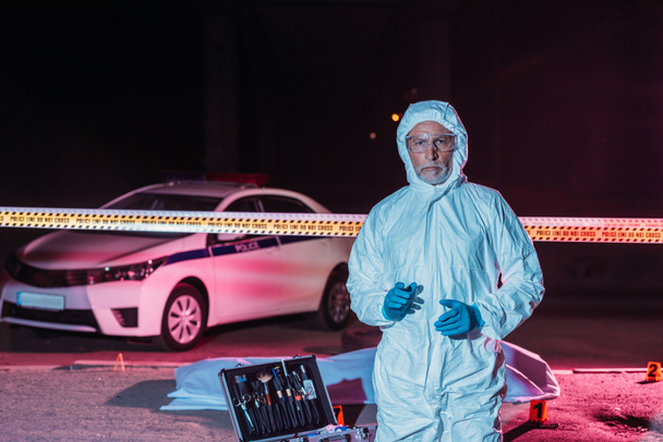 mature male criminologist in protective suit and mask looking at camera near crime scene with corpse in body bag  - Foto, Imagen