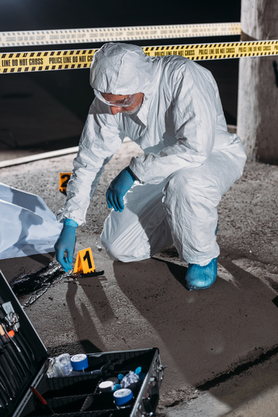 criminólogo masculino en traje de protección y guantes de látex recogiendo pruebas en la escena del crimen con el cadáver
  - Foto, Imagen