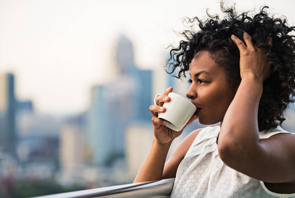 A close-up portrait of a woman standing on a terrace, drinking coffee. - Fotoğraf, Görsel