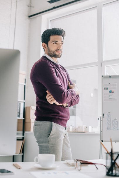 handsome designer in burgundy sweater standing with crossed arms and looking away in office  - Foto, Imagem