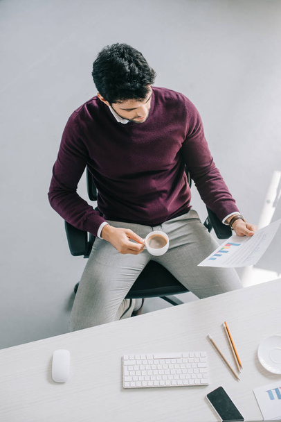 високий кут зору красивого дизайнера в бордовому светрі, що тримає чашку кави і читає документи в офісі
 - Фото, зображення