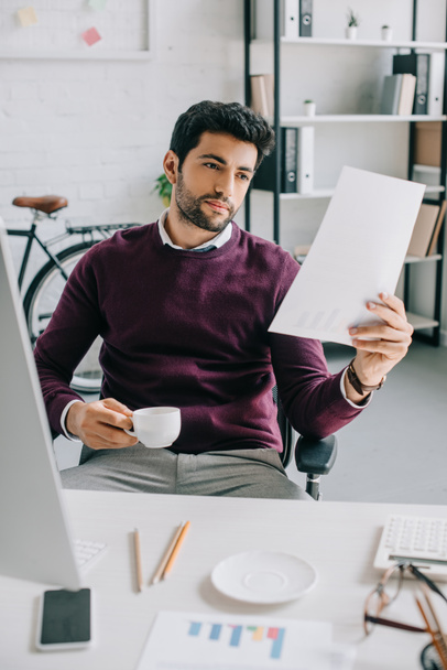 handsome businessman in burgundy sweater holding cup of coffee and reading documents in office - Foto, immagini
