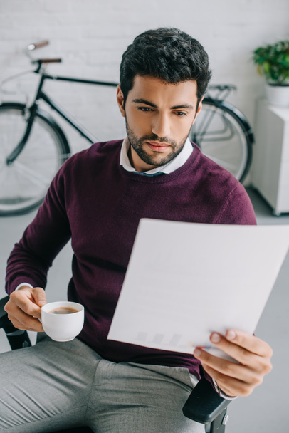 designer with bristle holding cup of coffee and reading documents in office - Photo, image