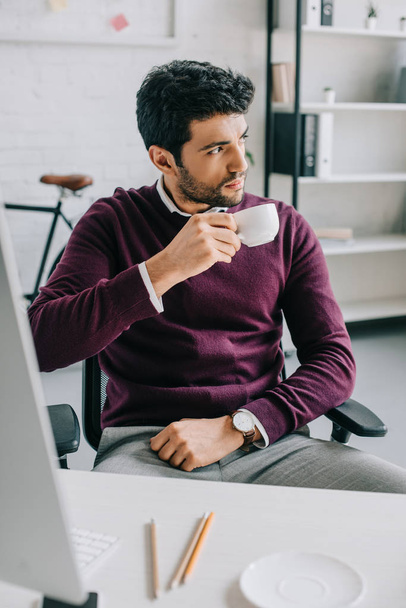 handsome businessman in burgundy sweater drinking coffee in office and looking away - Foto, Bild