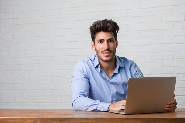 Mladí obchodní muž sedí a pracují na přenosném počítači s rukama na boky, stojící, uvolněný a usmívající se, velmi pozitivní a veselý - Fotografie, Obrázek