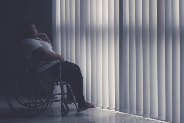 Side view of obese woman looks pensive while sitting in the wheelchair by the window. Shot in the hospital   - Photo, image