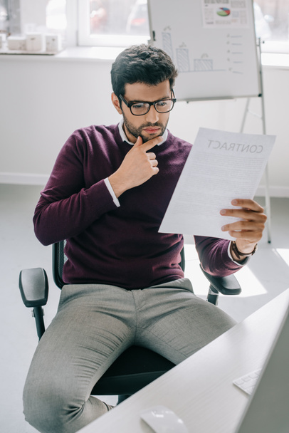 diseñador guapo pensativo en jersey de color burdeos sentado en la silla y la lectura de documentos en la oficina
 - Foto, imagen