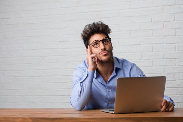 Un jeune homme d'affaires assis et travaillant sur un ordinateur portable réfléchissant et levant les yeux, confus à propos d'une idée, essaierait de trouver une solution
 - Photo, image
