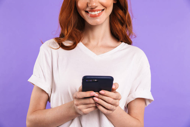 Cropped image of a smiling young girl standing isolated over violet background, using mobile phone - Fotografie, Obrázek