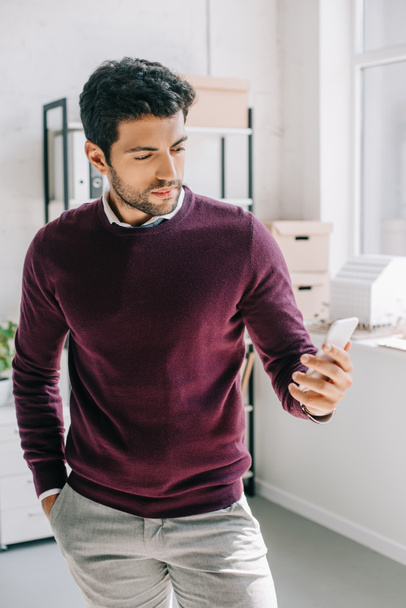 guapo diseñador en jersey de color burdeos mirando el teléfono inteligente en la oficina
  - Foto, imagen