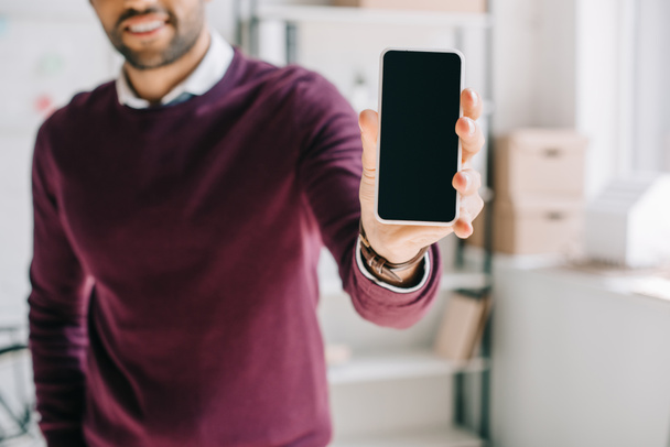 abgeschnittenes Bild eines lächelnden Designers im weinroten Pullover zeigt Smartphone mit leerem Bildschirm im Büro - Foto, Bild